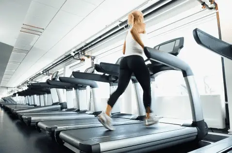Woman wearing a white top and black leggings and running on a treadmill near a lot of treadmills